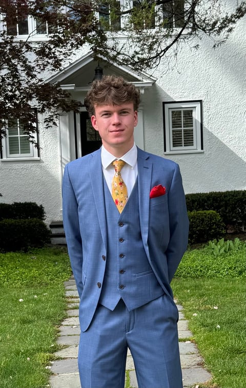 Young man in blue suit with yellow tie and red pocket square standing on walkway in front of white house