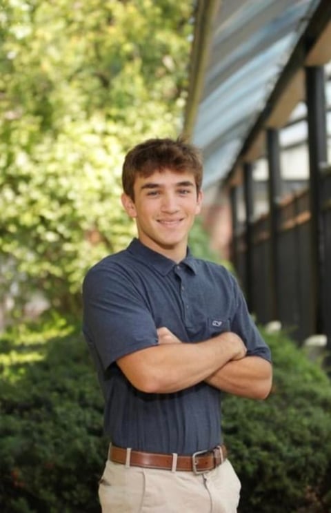Young man in navy polo shirt and khaki pants standing with crossed arms, smiling at camera with green foliage background